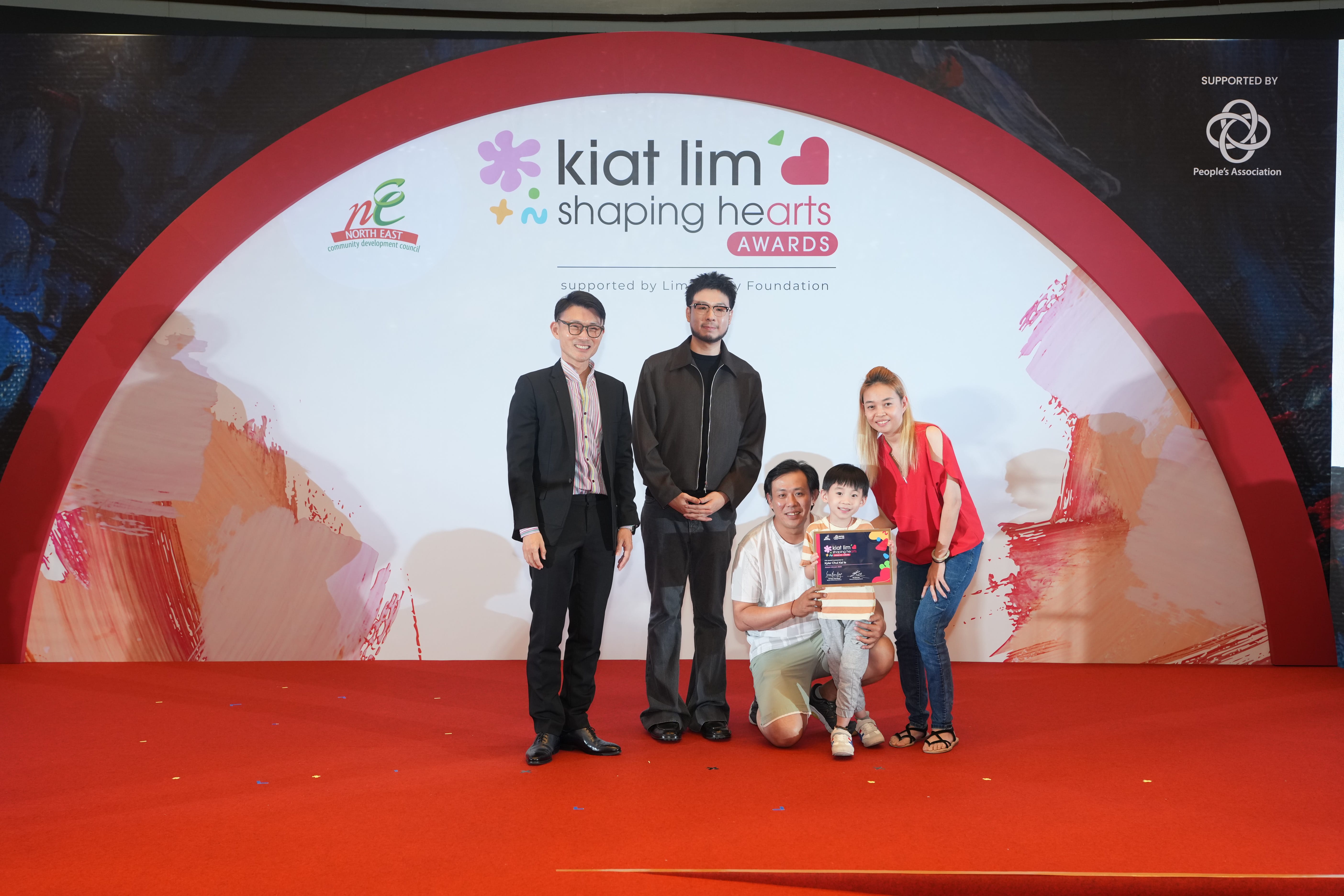 Award recipient standing on stage with the presenter, holding their certificate and posing for a group photo during the award ceremony, with a backdrop reading ‘Kiat Lim-Shaping Hearts Awards'.